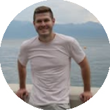 Smiling man in a light-colored t-shirt standing outdoors with mountains and water in the background.